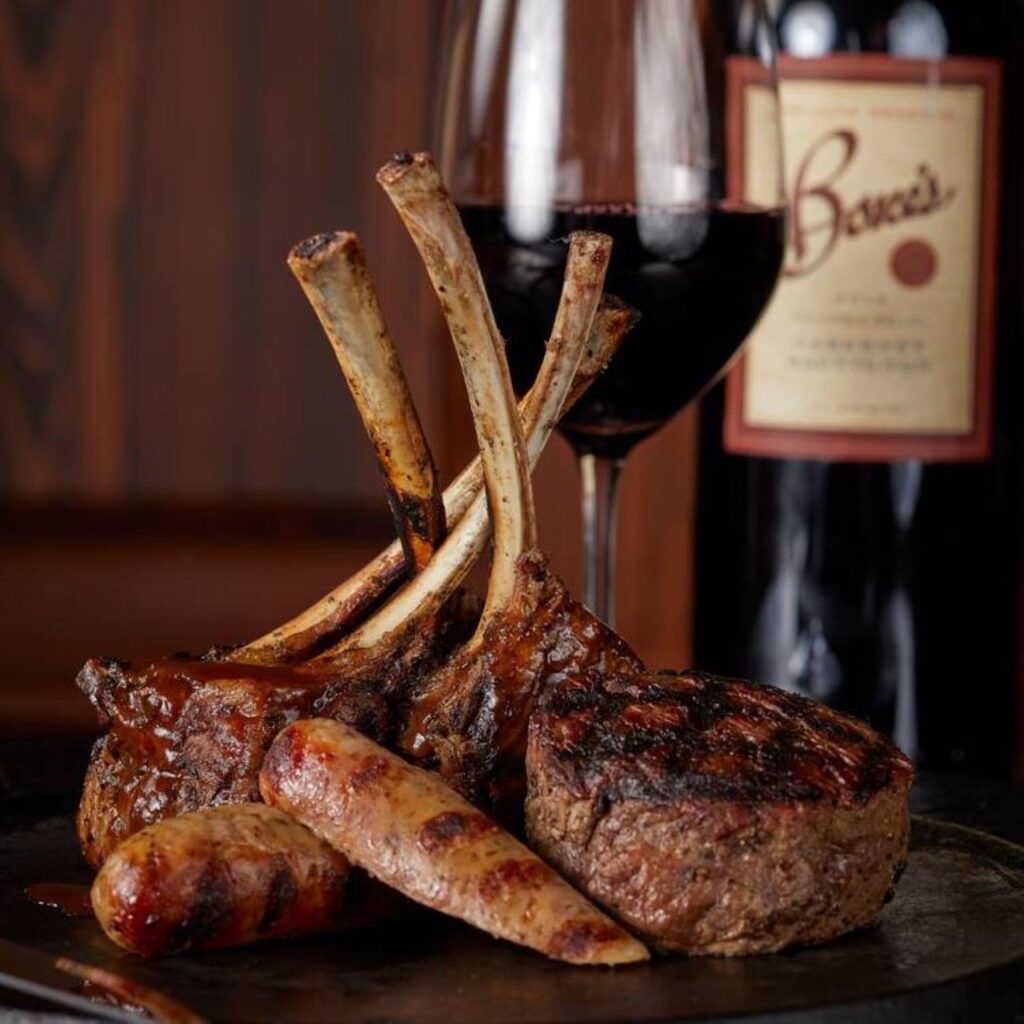 An image showcasing juicy Tomahawk steaks paired with beef sausage on a wooden cutting board. In the background, there's a glass of red wine next to an elegant bottle of wine