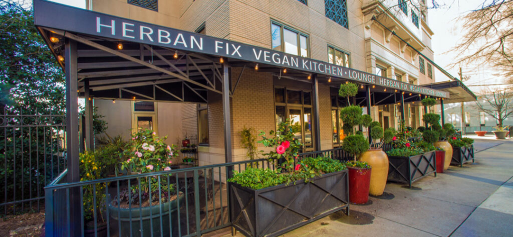 A picture of the outside of a restaurant on a street corner with plants and flowers