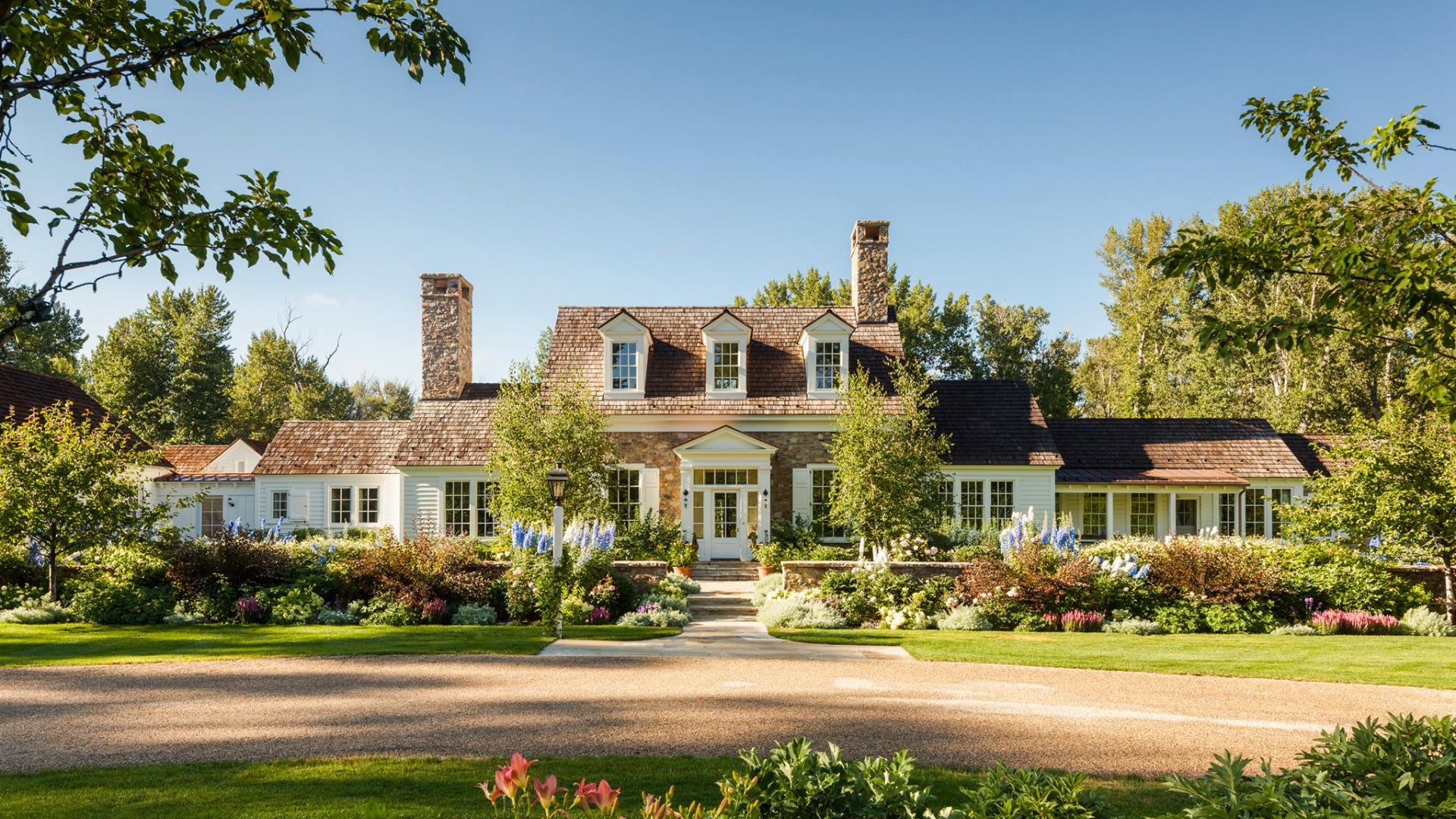 A grand cottage-style home nestled amidst lush foliage, adorned with vibrant plants, trees, and flowers that beautifully complement its picturesque surroundings.