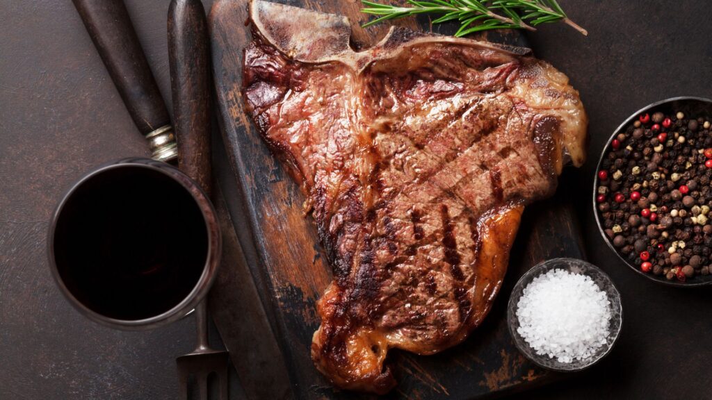 A nicely sized T-bone steak with sea salt and pepper on the side served on a rustic wooden board alongside a glass of wine.