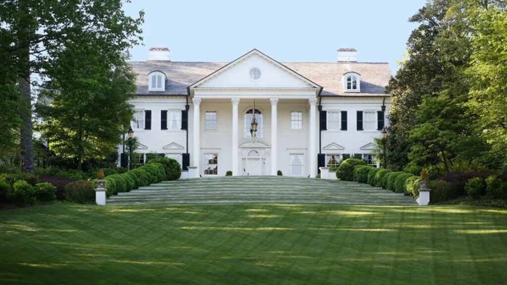 A magnificent home reminiscent of the White House, set amidst a lush green lawn and surrounded by towering trees, exuding grandeur and elegance.