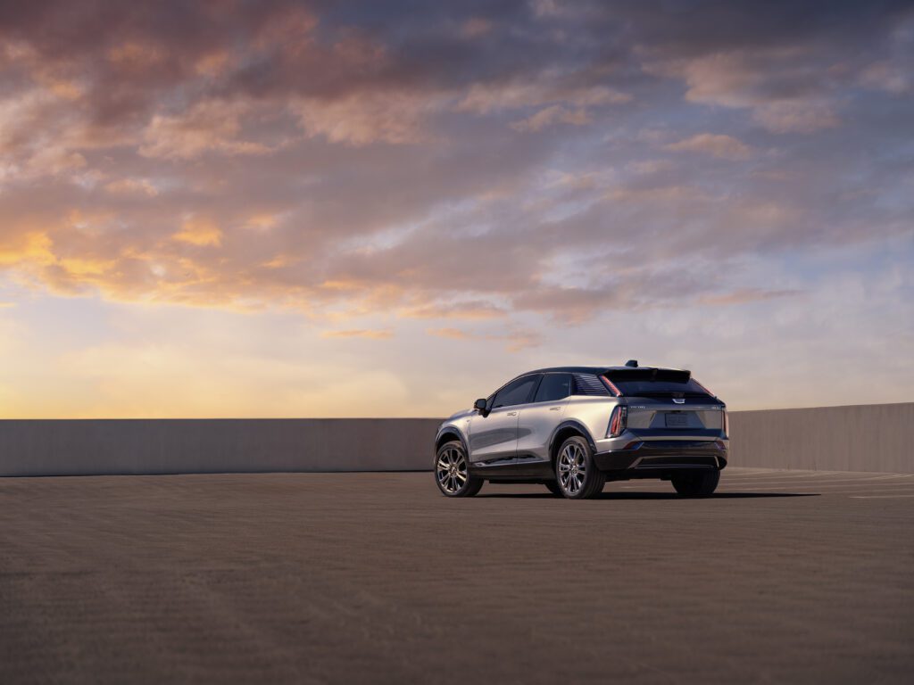 Rear three-quarter view of the Cadillac OPTIQ in Argent Silver Metallic parked against a dramatic evening sky backdrop.