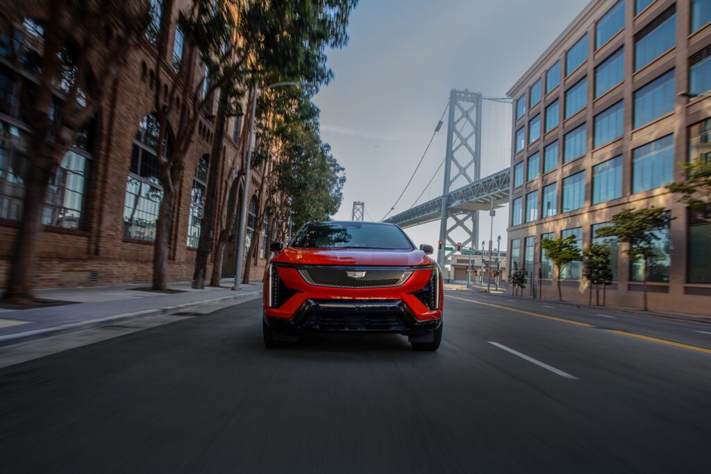 Front view of the Cadillac OPTIQ in Monarch Orange driving through an urban cityscape with the Bay Bridge in the background.