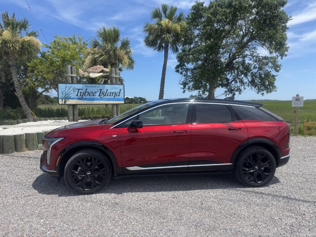 Red Cadillac OPTIQ parked in front of the Tybee Island welcome sign on a sunny day during a coastal road trip.

